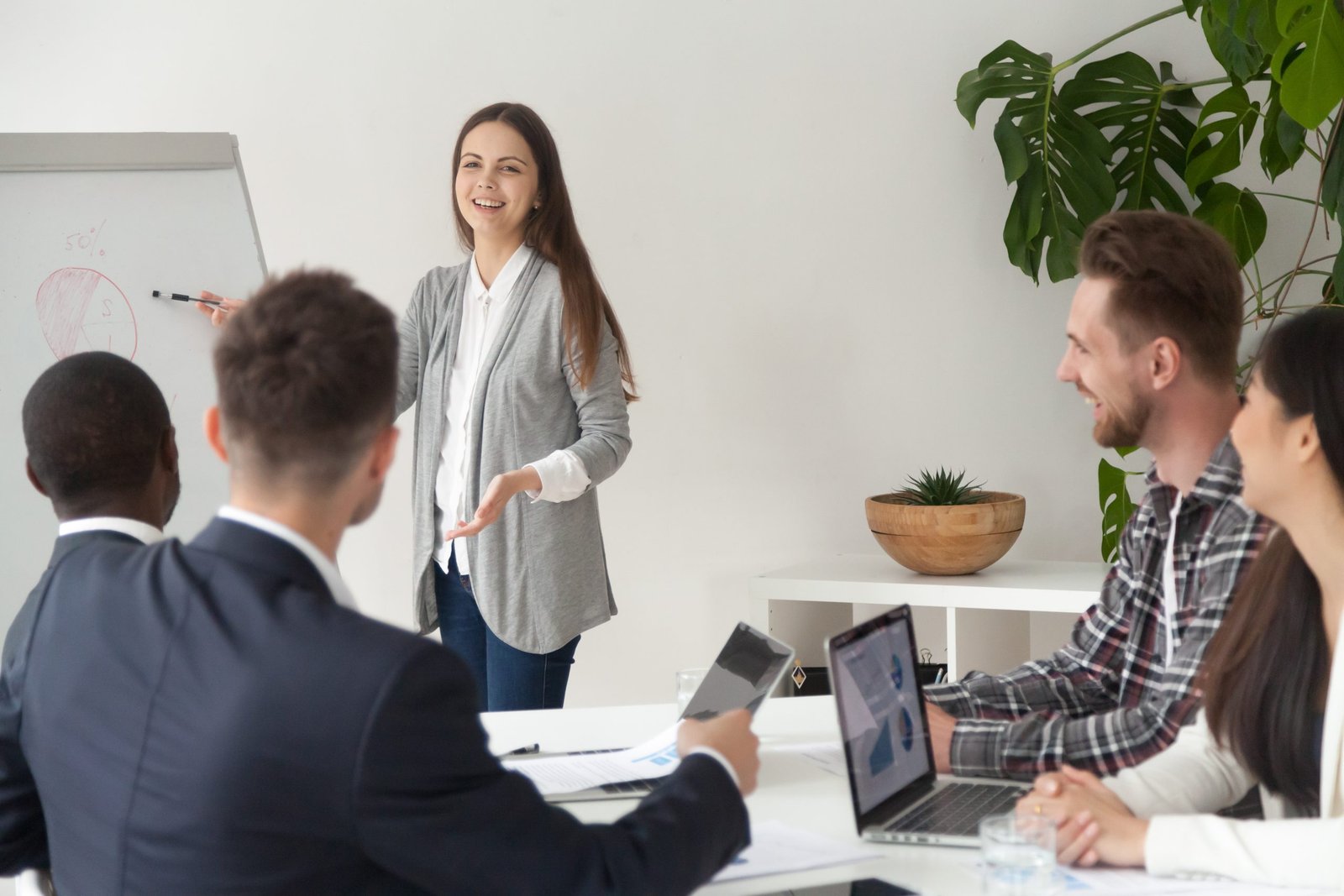 Smiling young employee or manager giving presentation working on flipchart in meeting room making business offer or reporting about project results, explaining new plan or idea to multi-ethnic team